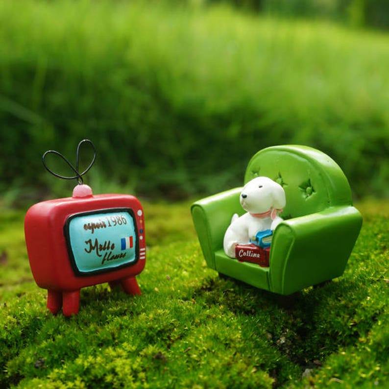 May include: A miniature red and white television with the text "expo 1986" and "Malle Fleurs" on the screen. A miniature green armchair with a white dog figurine sitting on it. The dog is holding a book with the text "Collins" on the cover.