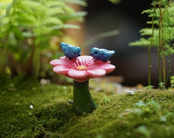 Op de afbeelding: Twee blauwe vogelbeeldjes die op een roze, bloemvormig vogelbad zitten, omringd door groen mos.