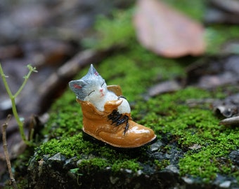 May include: A small, gray and white cat figurine sleeping inside a brown leather boot with black laces. The boot is sitting on a bed of green moss.