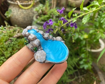 May include: A miniature waterfall and pond made of grey resin with a blue water feature. The waterfall and pond are sitting on a finger.