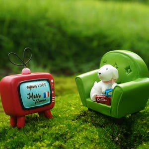 May include: A miniature red television with a white screen that says "espoir 1986" and "Mille fleurs" in French. A miniature white dog sits on a green miniature couch with a book in its paws.