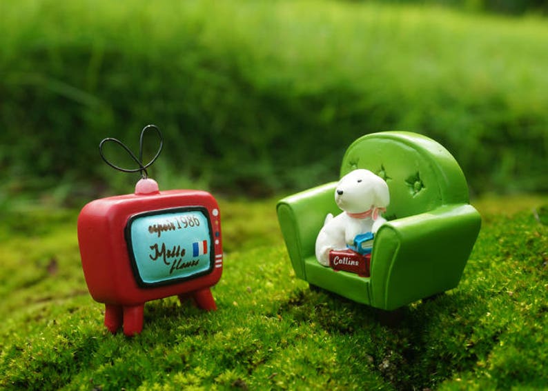 May include: A miniature red television with a white screen that says "espoir 1986" and "Mille fleurs" in French. A miniature white dog sits on a green miniature couch with a book in its paws.