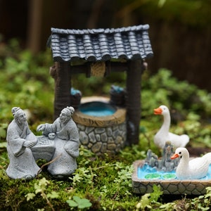 May include: A miniature scene with two gray figures playing a game, a small well with a gray roof, and two white ducks in a pond. The scene is set on a bed of green moss.
