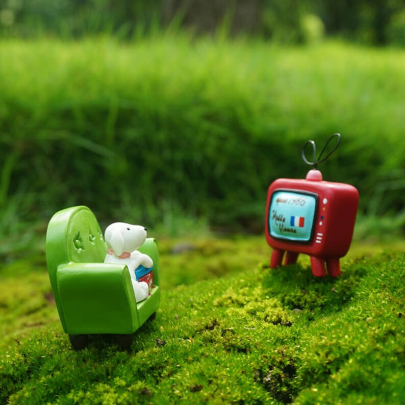 May include: A miniature green couch with a white dog figurine sitting on it, facing a red miniature television with a French flag on the screen. The scene is set on a bed of green moss.