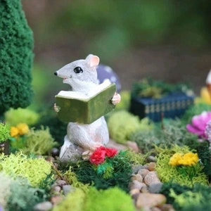 May include: A small gray mouse figurine sitting on a rock and holding a green book. The mouse is surrounded by green moss and small flowers.