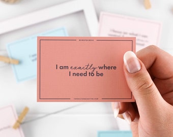 May include: A pink card with the text "I am exactly where I need to be" in dark gray font. The card has the word "MINDFULNESS" in smaller font at the top and "thedailygoalgetter.com" at the bottom.