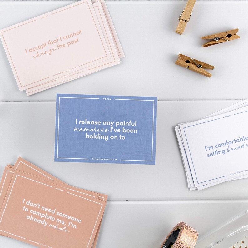 Puede incluir: Cuatro tarjetas de afirmaci&oacute;n con mensajes positivos impresos en ellas. Las tarjetas son de color rosa, azul y melocot&oacute;n. Las tarjetas est&aacute;n sobre una superficie de madera blanca con pinzas de madera y un rollo de cinta.