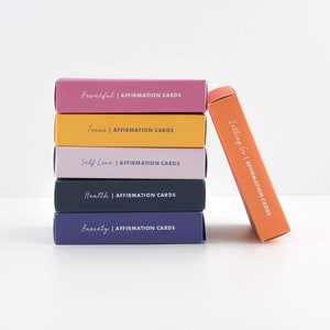 K&ouml;nnte beinhalten: Ein Stapel aus sechs bunten Boxen mit den Worten "Affirmation Cards" auf jeder Box. Die Boxen sind mit verschiedenen Themen beschriftet, darunter "Powerful", "Teens", "Self Love", "Health", "Anxiety" und "Letting Go".