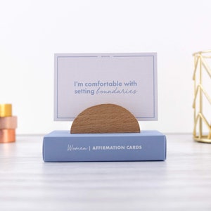 Puede incluir: Una caja azul con el texto "Women | Affirmation Cards". Una tarjeta blanca con el texto "I'm comfortable with setting boundaries" se muestra en un soporte de madera.