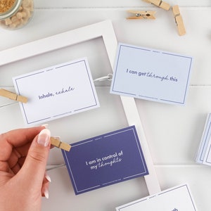 K&ouml;nnte beinhalten: Ein Satz Affirmationskarten mit positiven Botschaften zur Bew&auml;ltigung von Angstzust&auml;nden. Die Karten sind auf wei&szlig;em und violettem Papier mit schwarzem Text gedruckt. Die Karten werden in einem wei&szlig;en Rahmen mit Holz-W&auml;scheklammern pr&auml;sentiert. Der Text auf den Karten lautet: "Einatmen, Ausatmen", "Ich kann das schaffen", und "Ich habe meine Gedanken im Griff".
