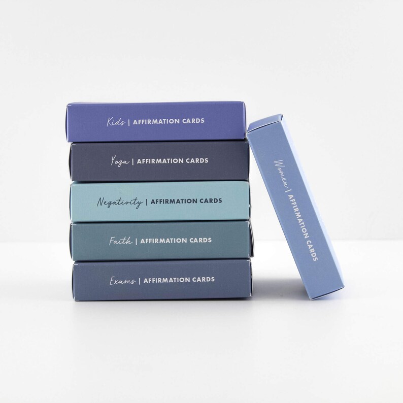Puede incluir: Una pila de seis cajas azules y grises con el texto "AFFIRMATION CARDS" en cada caja. Las cajas est&aacute;n etiquetadas con diferentes temas, que incluyen "Kids", "Yoga", "Negativity", "Faith", "Exams" y "Women".