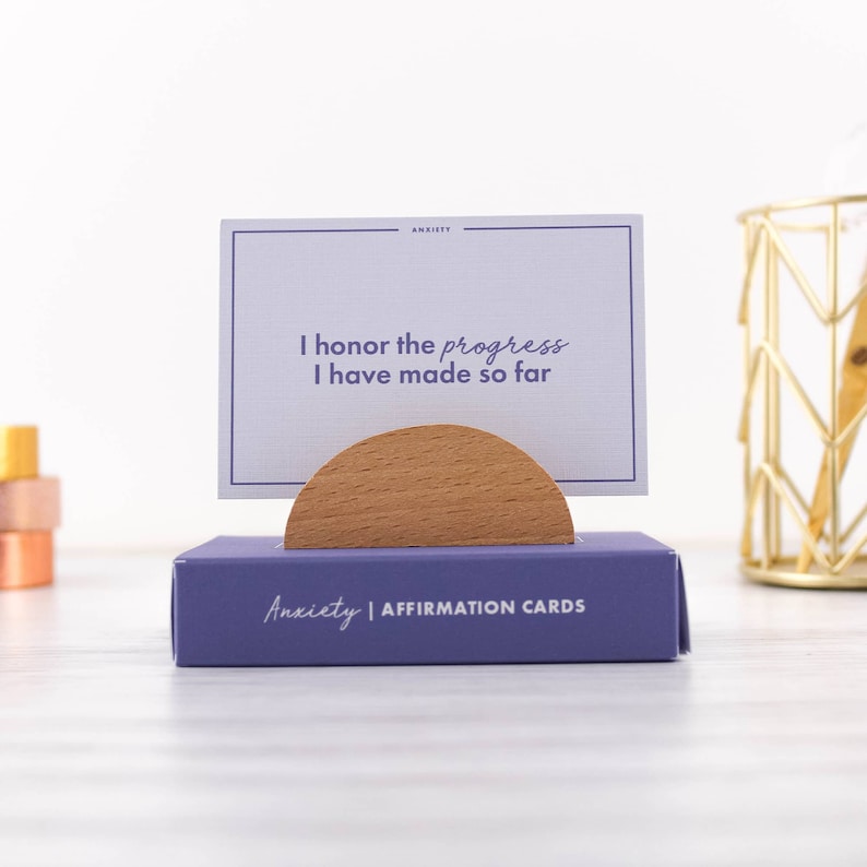 K&ouml;nnte beinhalten: Lilafarbener Kasten mit dem Text "Anxiety | Affirmation Cards" auf der Vorderseite. Ein Holzst&auml;nder h&auml;lt eine Karte mit dem Text "Ich ehre den Fortschritt, den ich bisher gemacht habe".
