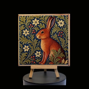 May include: A framed artwork depicting a brown hare amidst a floral design in green, cream, and red on a dark blue background. The artwork is displayed on a small wooden easel. The artwork is square in shape.