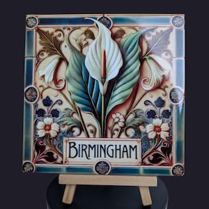 May include: A ceramic tile with a floral design featuring white calla lilies and green leaves. The tile has a blue and white border and the word "BIRMINGHAM" in a gold banner.