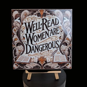 May include: Square ceramic tile with the text "Well Read Women Are Dangerous" in a decorative font. The tile features floral and scroll designs in brown, white, and gold. Two open books and a quill are also depicted.