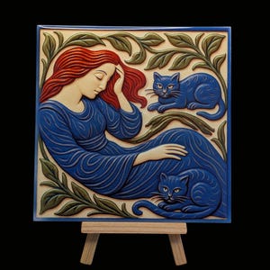 May include: A square relief artwork depicting a woman with red hair in a blue dress, with two blue cats. Green leaves and branches surround the figures. The artwork is displayed on a small wooden easel.