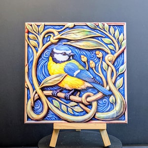 May include: A square tile depicting a blue, yellow, and white bird on a branch, set against a blue backdrop with gold botanical details. The artwork is presented on a small wooden easel.