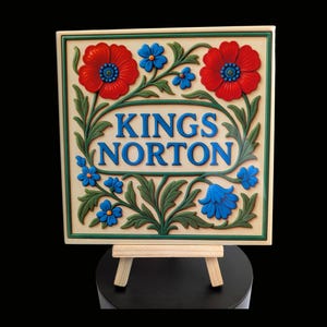 May include: A cream-coloured sign featuring the words "KINGS NORTON" in blue, framed by red poppies, blue flowers, and green foliage. The sign is displayed on a small wooden easel.
