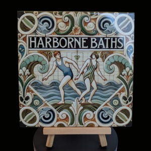 May include: A vintage ceramic tile with an ornate border and a scene of two people in swimming costumes swimming in the sea. The tile reads "Harborne Baths".