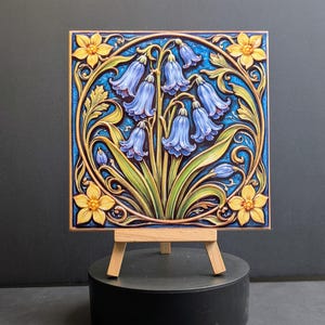 May include: A square tile with a relief design of bluebell flowers and yellow blossoms, set against a blue backdrop. The tile is presented on a small wooden easel. The design showcases the intricate floral details and colour scheme.