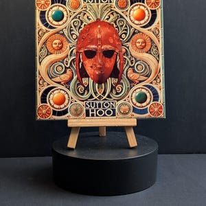 May include: Square tile with a red helmet illustration and the text "SUTTON HOO". The design incorporates intricate patterns, sun-like faces, and bird motifs, set against a dark blue background. The tile is displayed on a small wooden easel.