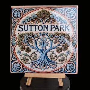 May include: A ceramic tile featuring a blue and green tree design with the words "Sutton Park" in a white banner. The tile is displayed on a small wooden easel.