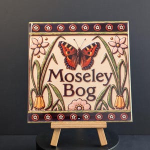 May include: A square ceramic tile featuring the text Moseley Bog in dark brown lettering. The design includes a central orange and black butterfly, light pink flowers, and green foliage. The top and bottom borders are dark purple with small white flowers.