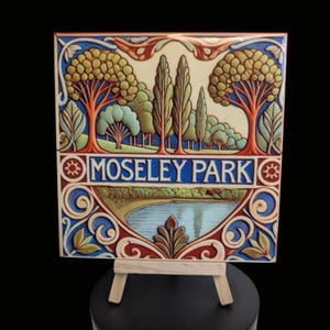 May include: A ceramic tile featuring a colourful illustration of a park scene. The tile depicts a pond, trees, and the text "Moseley Park". The tile is displayed on a small wooden easel.
