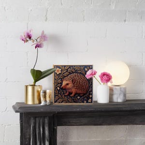 May include: A decorative square art print featuring a hedgehog in a floral design, displayed on a dark mantel. The scene includes a gold planter with an orchid, a small vase with pink flowers, and a marble-based lamp.