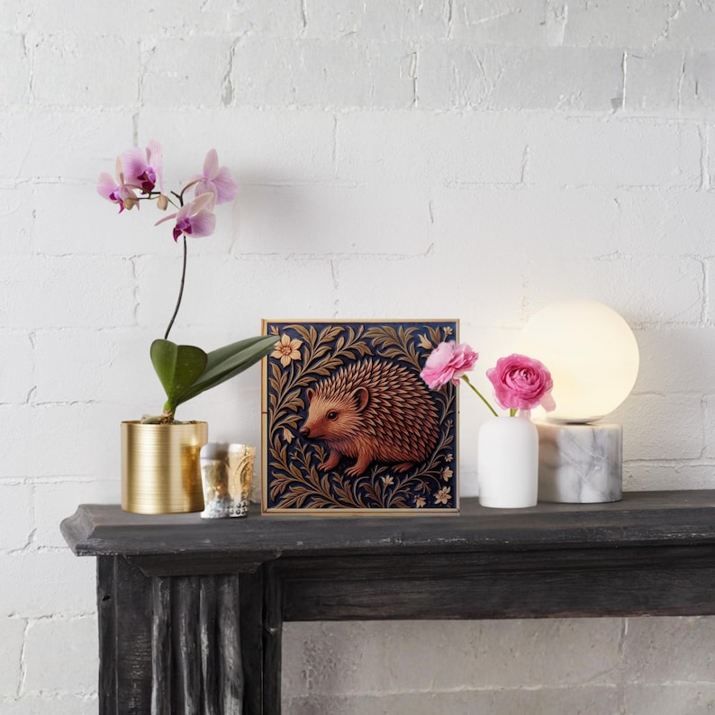 May include: A decorative square art print featuring a hedgehog in a floral design, displayed on a dark mantel. The scene includes a gold planter with an orchid, a small vase with pink flowers, and a marble-based lamp.