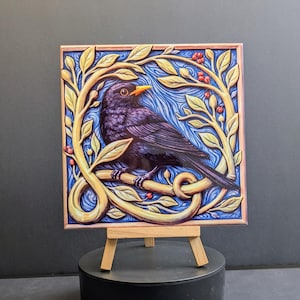 May include: Square art print depicting a blackbird on a branch, framed by gold-coloured leaves and red berries. The background is blue with a textured design. The artwork is presented on a small wooden easel.