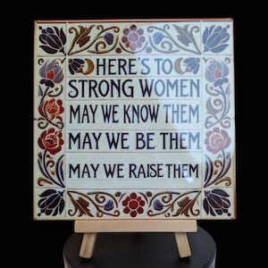 May include: A white ceramic tile with a floral border and the text "HERE'S TO STRONG WOMEN MAY WE KNOW THEM MAY WE BE THEM MAY WE RAISE THEM". The tile is displayed on a small wooden easel.