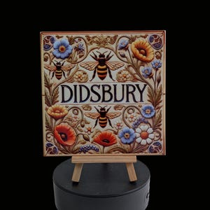 May include: Square ceramic tile with the word "DIDSBURY" in a decorative font, framed by flowers and bees. The tile is displayed on a small wooden easel, showcasing a vintage, floral design. The tile is likely a decorative item.