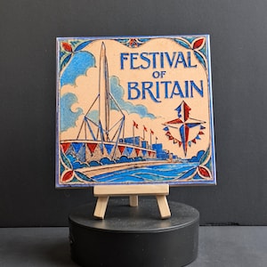 May include: A square ceramic tile with the words "FESTIVAL OF BRITAIN". The tile shows a building with a spire, flags, and a compass rose. The tile is displayed on a small wooden easel. The design is framed with a decorative border.