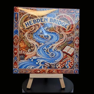 May include: A decorative tile with a blue and gold design featuring a river flowing through a landscape. The tile is framed with a decorative border and has the words "Hebden Bridge" written in a stylised font. The tile is displayed on a wooden easel.