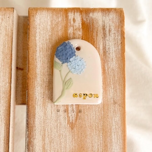 Puede incluir: Una etiqueta crema en forma de arco con un diseño floral que presenta hortensias azules y la palabra "aspen" en letras doradas. La etiqueta tiene un pequeño agujero en la parte superior y está sobre un fondo de madera.