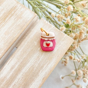 May include: A small pink ceramic charm shaped like a jar of strawberry jam with a gold loop for hanging.