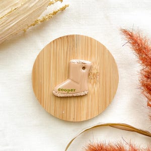 May include: A light brown ceramic boot charm with the name "cooper" written in gold lettering.