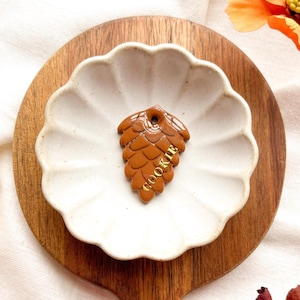 May include: A brown cookie cutter in the shape of a pinecone, with the word "COOKIE" in gold lettering, rests on a white scalloped dish. The dish sits on a wooden cutting board, with a white cloth and orange flowers in the background.
