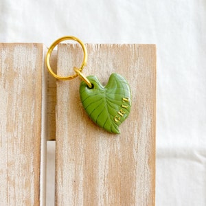 Könnte beinhalten: Ein grüner, blattartiger Schlüsselanhänger mit einem goldenen Ring und dem Wort "GRIT" in goldenen Buchstaben.