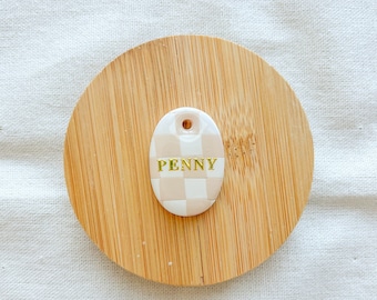 May include: A white ceramic oval tag with a light brown and white checkered pattern and the word "PENNY" in gold lettering.