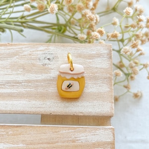 May include: A small, yellow honey jar charm with a white lid and a black and yellow bee design. The charm is attached to a gold jump ring.