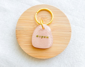 May include: A pink, textured, oval-shaped keychain with a gold ring and the word "aspen" written in gold lettering.