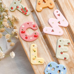 May include: A collection of colorful, decorative letter charms. Each letter is a different pastel color and adorned with miniature fruit and flower designs. The letters are arranged on a wooden surface, creating a charming, handcrafted aesthetic.
