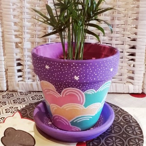 Op de afbeelding: Paarse geverfde terracotta pot met een witte gespikkelde achtergrond en een regenboogwolk ontwerp. De pot heeft een bijpassende schotel.