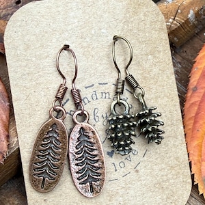 May include: Handmade earrings featuring copper-toned oval pendants with etched tree designs and bronze-toned pine cone charms. The earrings are displayed on a craft paper card with the text "handmade by love".