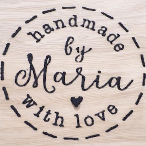 May include: A wooden surface with the words "handmade by Maria with love" in black script. The text is arranged in a circle, with a small heart symbol. The background is a light wood grain.