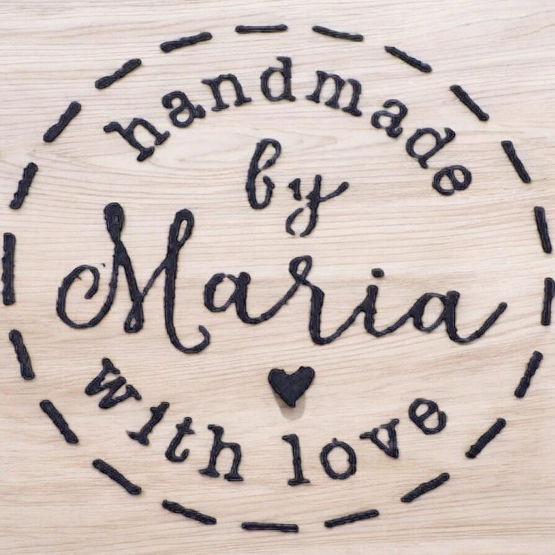 May include: A wooden surface with the words "handmade by Maria with love" in black script. The text is arranged in a circle, with a small heart symbol. The background is a light wood grain.