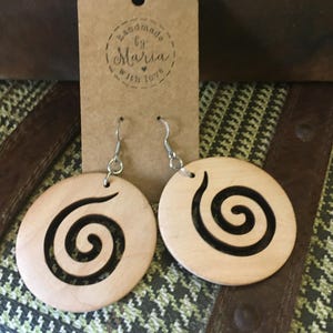 May include: A pair of wooden earrings with a spiral design. The earrings are circular, light-colored wood with a black spiral cut-out. The earrings hang from silver-colored hooks. A brown tag with the text "handmade by Maria with love" is behind the earrings.