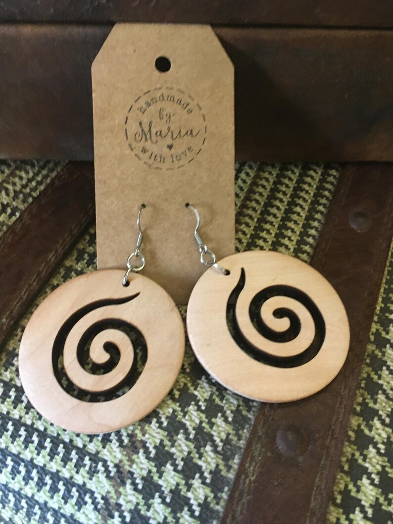 May include: A pair of wooden earrings with a spiral design. The earrings are circular, light-colored wood with a black spiral cut-out. The earrings hang from silver-colored hooks. A brown tag with the text "handmade by Maria with love" is behind the earrings.